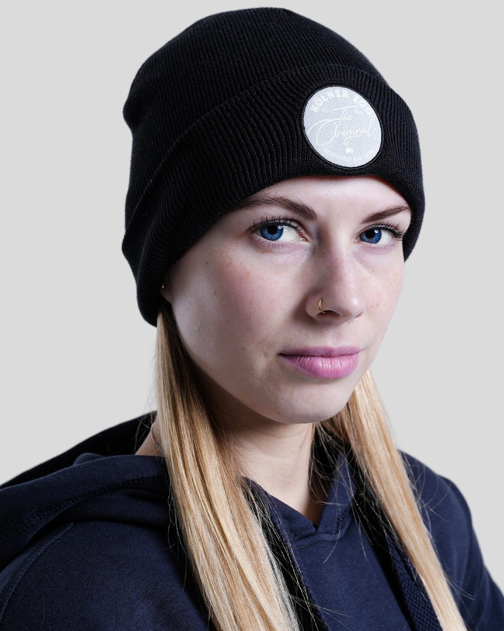Schwarze Strickmütze mit breitem Umschlag, getragen von einer Person mit blonden Haaren. Auf der Vorderseite ist ein runder, heller Aufnäher mit der Aufschrift ‚Kölner Zoo – The Original‘ in Schreibschrift angebracht.