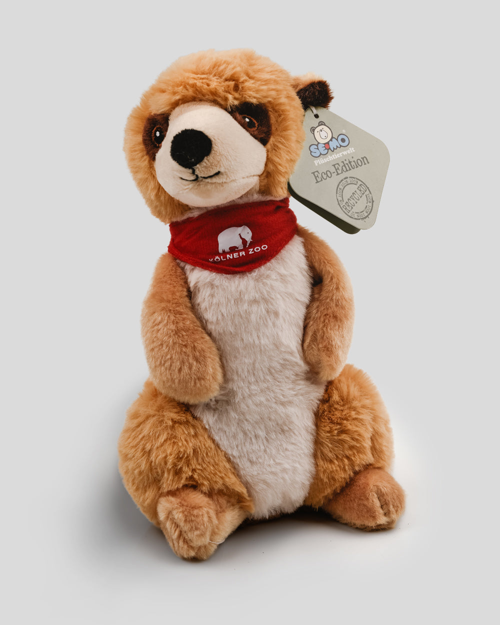 Plüsch-Erdmännchen in aufrechter Haltung mit weichem, braun-beigem Fell. Das Kuscheltier trägt ein rotes Halstuch mit weißem Elefantensymbol und dem Schriftzug ‚Kölner Zoo‘. An einem Ohr ist ein Etikett mit der Aufschrift ‚SEMO Plüschverwert – Eco-Edition‘ und dem Hinweis auf recyceltes Material befestigt.