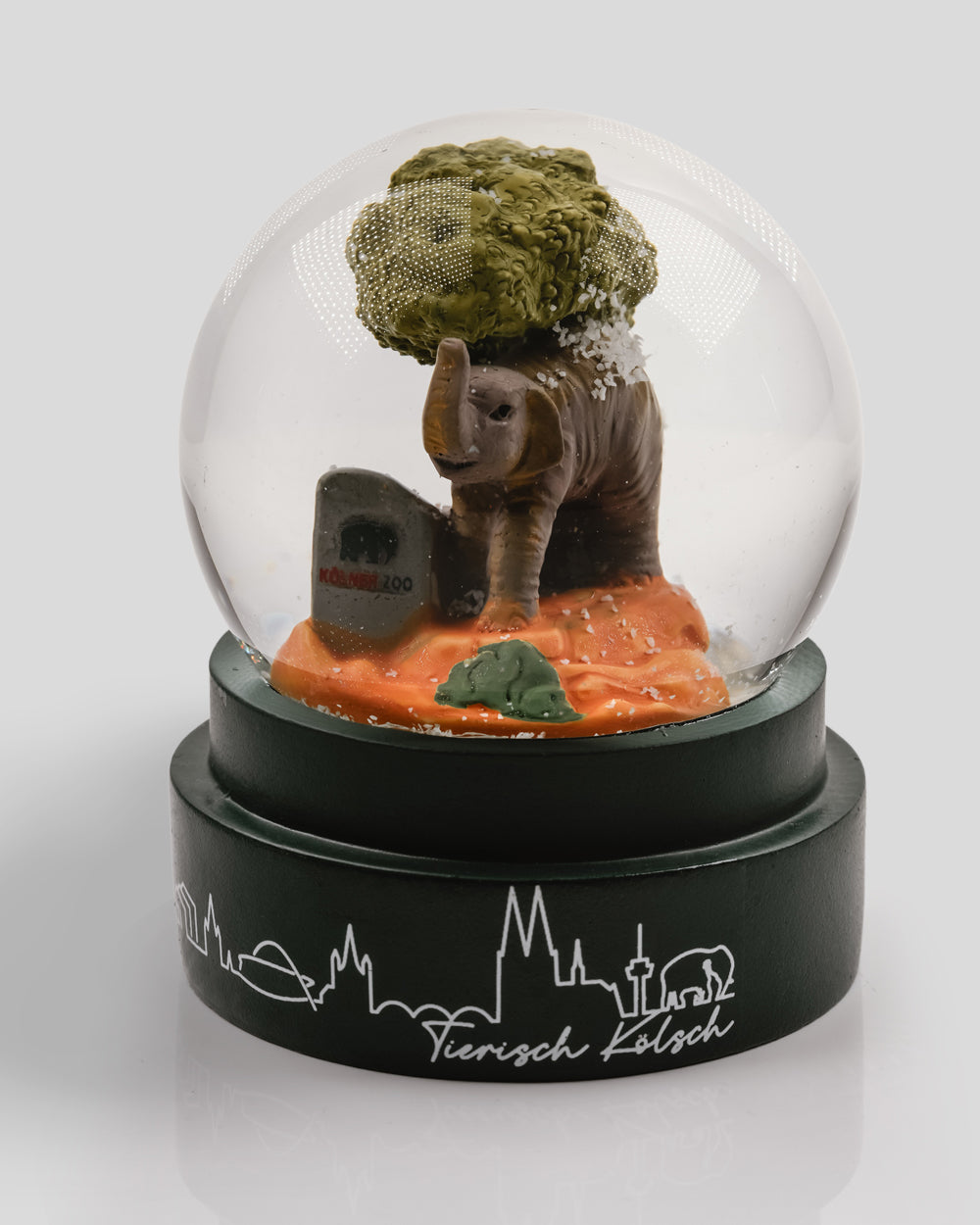 Dekorative Schneekugel mit Elefantenfigur, grünem Baum und einem kleinen Stein mit ‚Kölner Zoo‘-Logo im Inneren. Die Figur steht auf orangem Boden, Schneeflocken wirbeln beim Schütteln umher. Der Sockel ist dunkelgrün mit weißer Kölner Skyline und dem Schriftzug ‚Tierisch Kölsch‘ verziert.