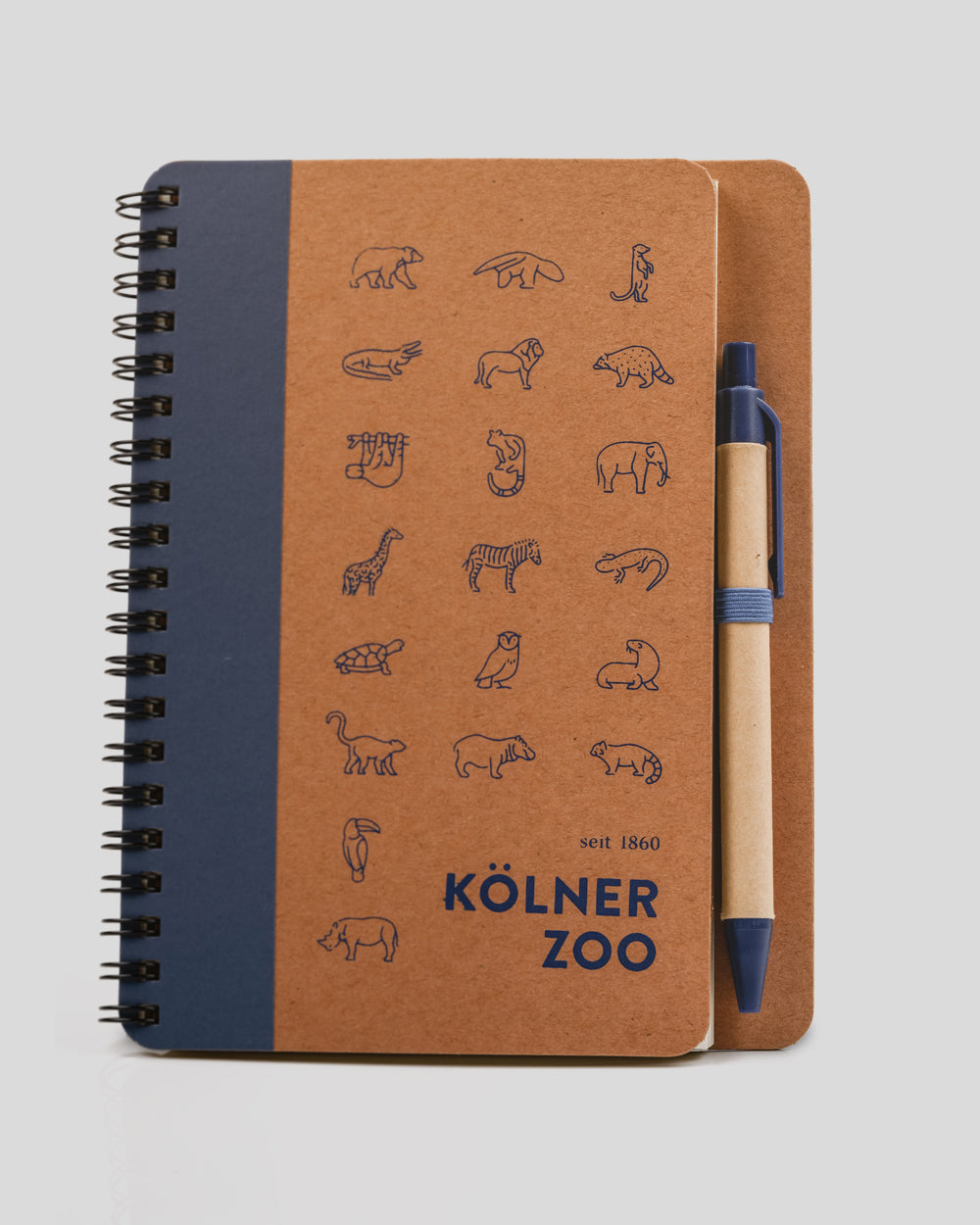 Notizbuch mit braunem Recycling-Kartonumschlag und schwarzer Spiralbindung. Auf dem Cover sind 20 dunkelblaue Tierillustrationen sowie der Schriftzug ‚Kölner Zoo – seit 1860‘ aufgedruckt. An der Seite befindet sich ein passender Stift in Natur-Optik mit blauer Kappe in einer elastischen Halterung.