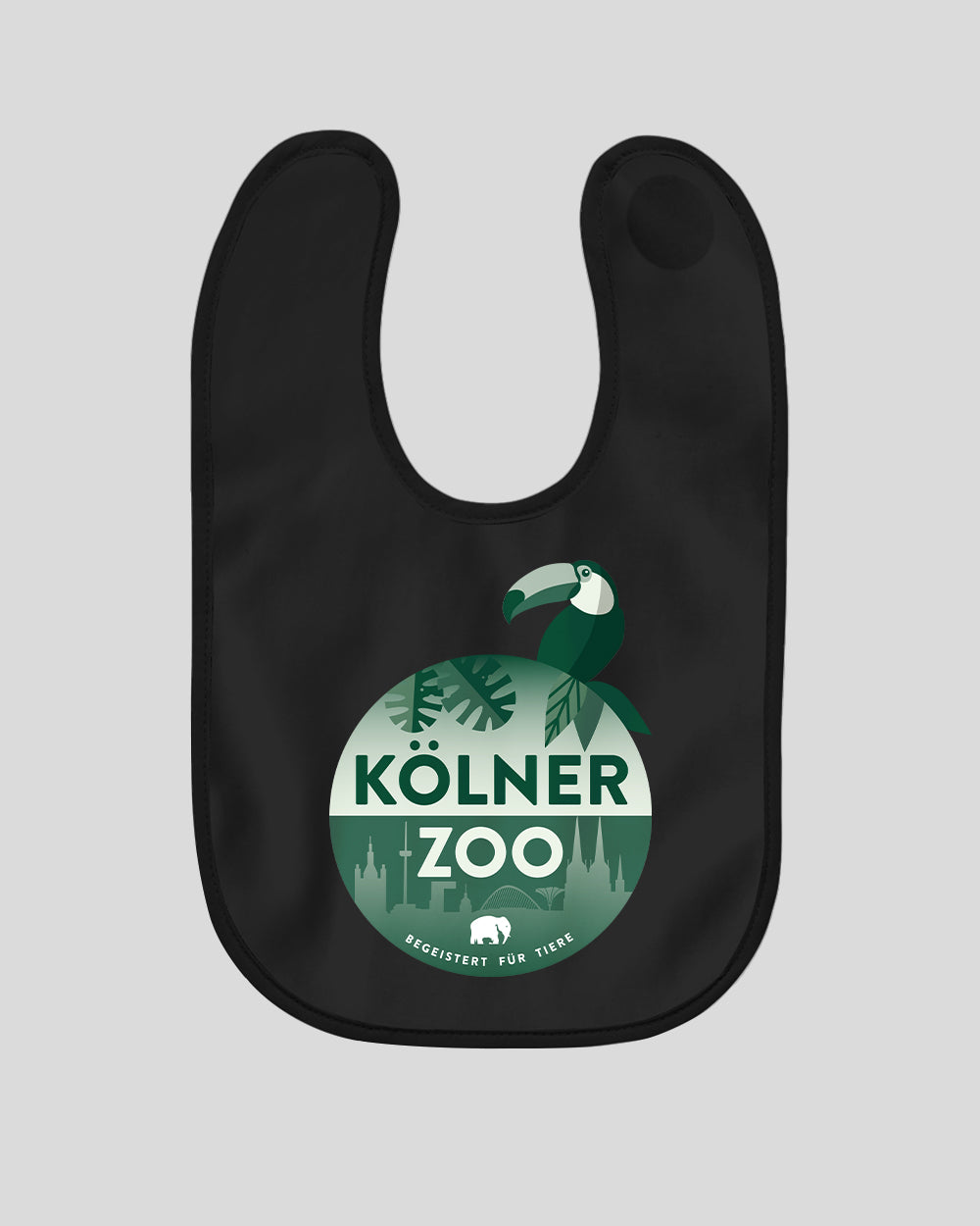Ein schwarzes Lätzchen mit einem runden, grün gestalteten Logo in der Mitte. Auf dem Logo steht „Kölner Zoo“ über einer stilisierten Skyline von Köln. Darüber sind tropische Blätter und ein grüner Tukan abgebildet. Unten im Kreis steht der Slogan „Begeistert für Tiere“ mit einem kleinen weißen Elefantensymbol.