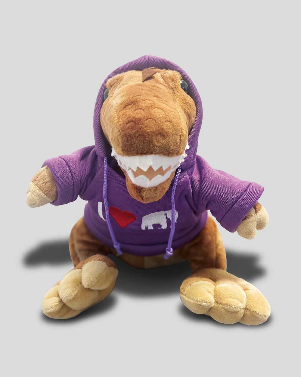 Plüsch-Dino mit lilafarbenen Hoodie, darauf ein der Stick "I rotes herzsymbol KölnerZoo Logo". Der Dino hat große weiße Zähne, schwarze Knopfaugen und die Kaputze über den Kopf gezogen