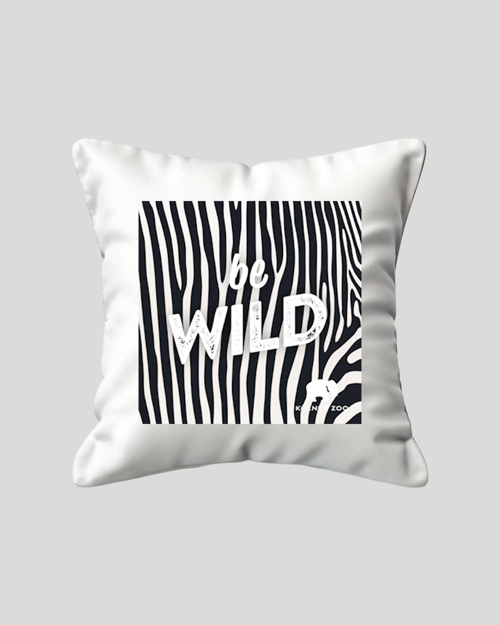 Weißes Zierkissen mit quadratischem Aufdruck im schwarz-weißen Zebramuster. In der Mitte steht der weiße Schriftzug ‚be WILD‘ in markanter Typografie. Unten rechts befindet sich ein kleines Elefantensymbol mit dem Text ‚Kölner Zoo‘.